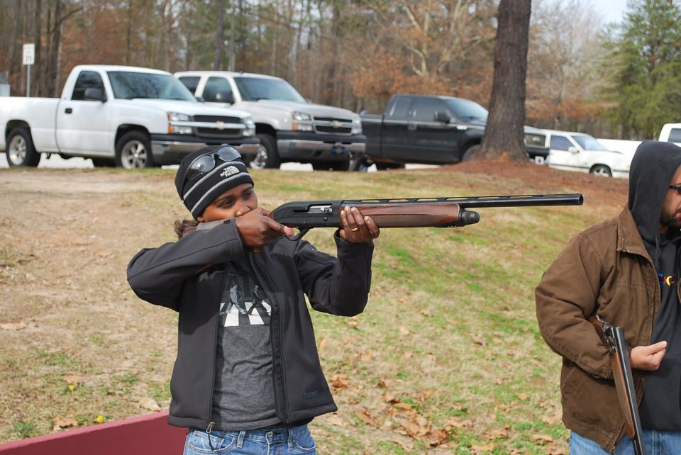 Person aims shotgun, another person stands nearby with a shotgun. Pickup trucks are parked in the background