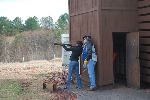 1979 Ford F-150 - A woman aims a shotgun while an instructor watches at a shooting range, with a wooden structure and trees in the background.