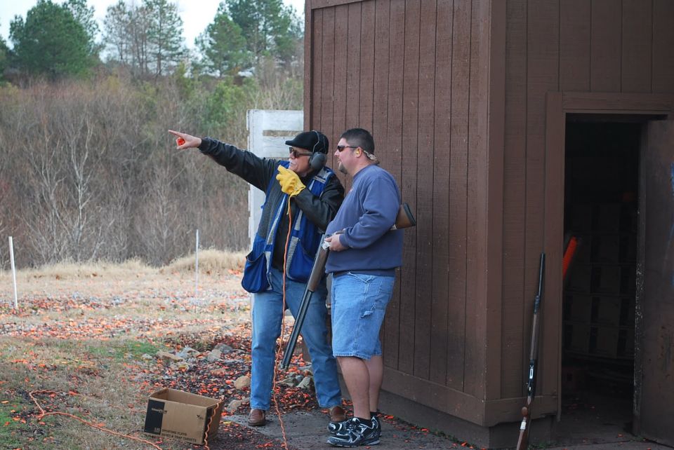 Man in blue vest points, man in blue sweatshirt holds shotgun at shooting range