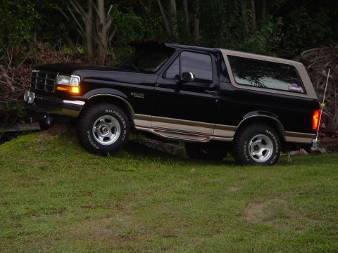 1996 Ford Bronco - A black 1996 Ford Bronco with tan accents is parked on a grassy slope, surrounded by trees.