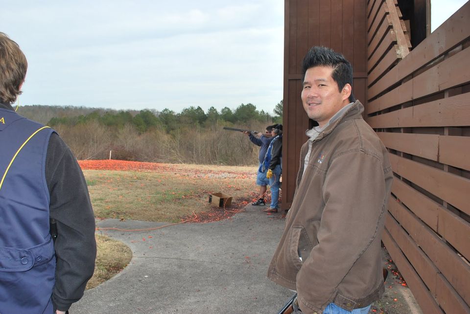 Man in brown jacket smiles near wooden structure at outdoor shooting range with clay targets scattered