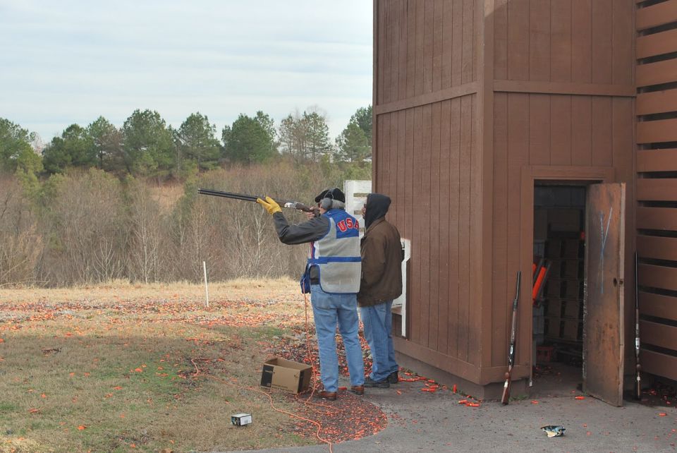 Two people at a clay shooting range, one aiming a shotgun. A wooden structure is on the right