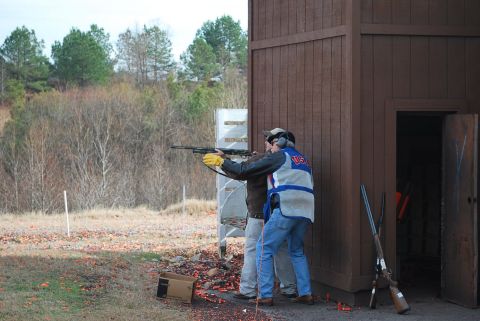 1979 Ford F-150 - A shooting instructor guides a student at a clay pigeon range with a wooden structure and trees in the background.