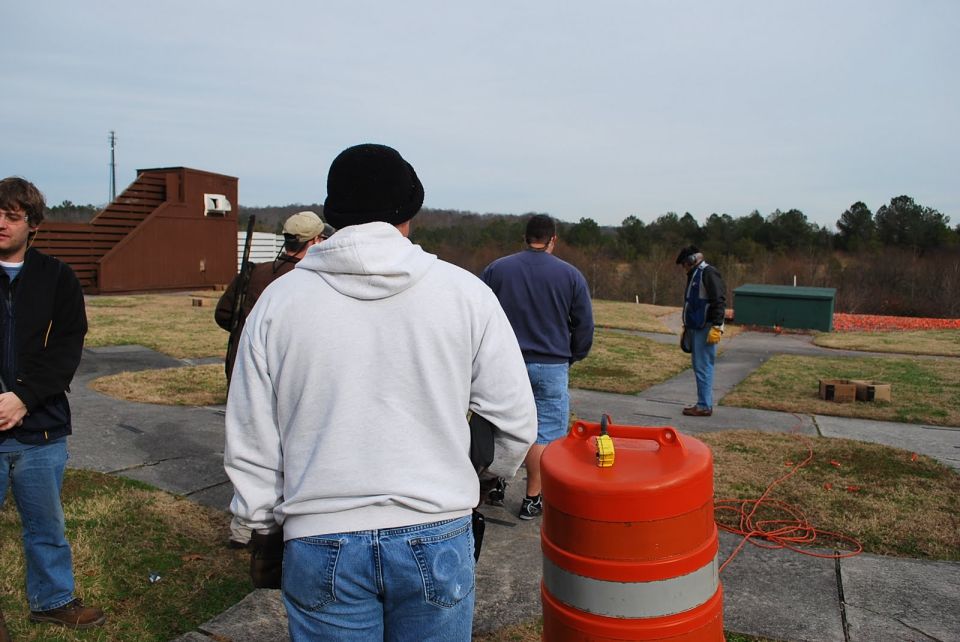 People stand on a paved area at an outdoor shooting range with clay targets and structures in the background
