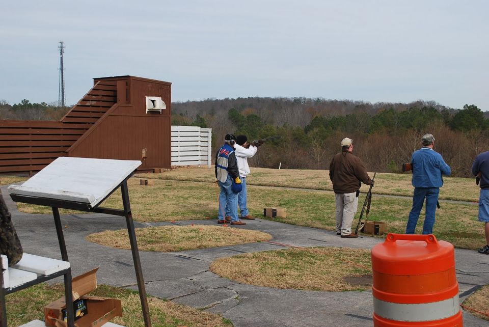 Outdoor shooting range with people aiming shotguns, a wooden structure, and a cell tower in the distance