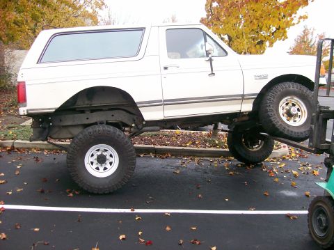 1989 Ford Bronco - A white Ford Bronco is lifted by a forklift, revealing its lifted suspension and large tires.