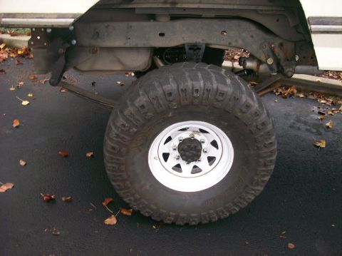 1989 Ford Bronco - The rear wheel and suspension of a vehicle are shown, with a prominent off-road tire and leaf-strewn pavement.