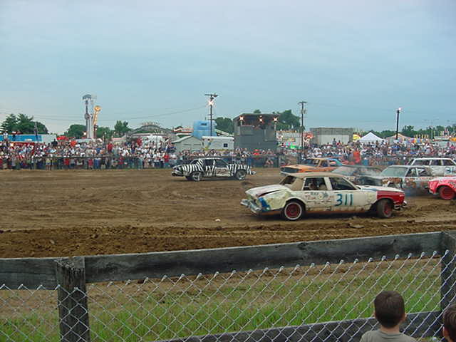 White demolition derby sedan #311 with crushed front end on dirt track, other derby cars and crowd behind