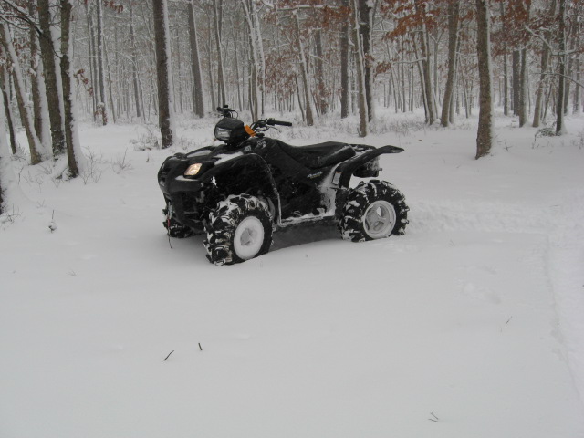 Black ATV parked in snow on a wooded trail, snow covering its tires and racks