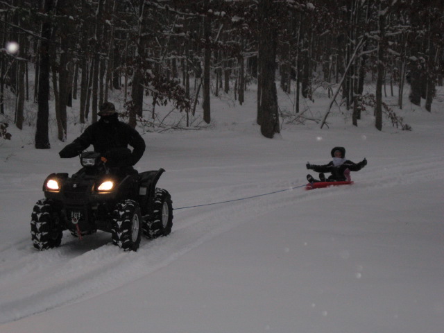Black Suzuki ATV with headlights on towing a child on a red sled through snowy woods