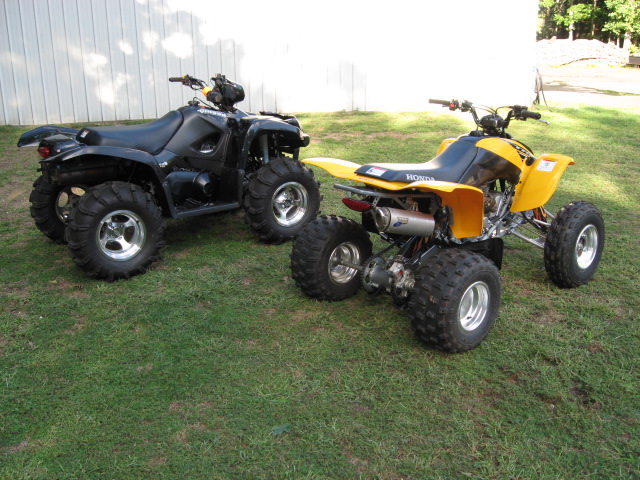 Black Suzuki Vinson ATV and yellow Honda sport quad parked on grass by a white building
