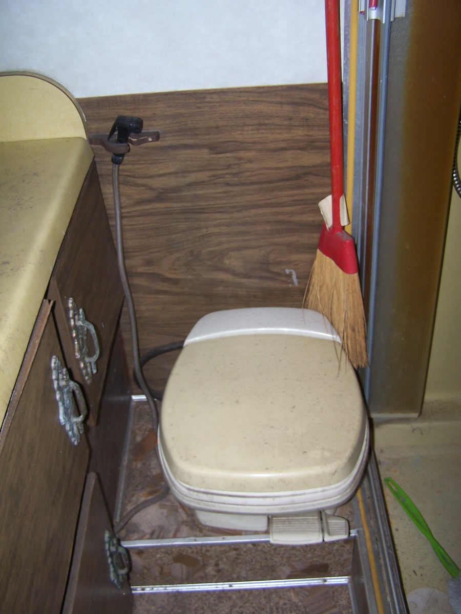 Tan cassette toilet inside a wood-paneled RV bathroom with a broom