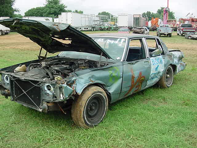Crashed teal sedan with open damaged hood and spray paint, parked on grass at a fairground