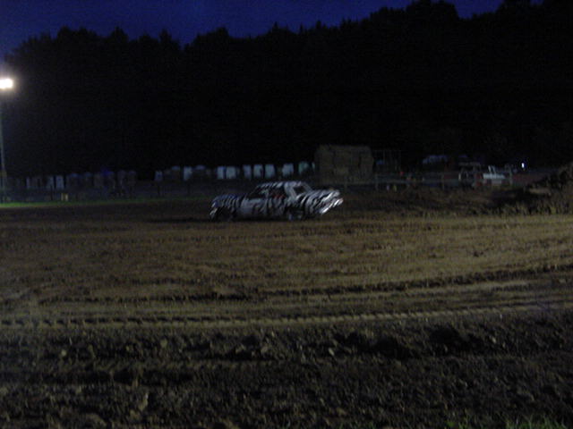 Painted demolition derby car driving on a dirt track at dusk under stadium lights