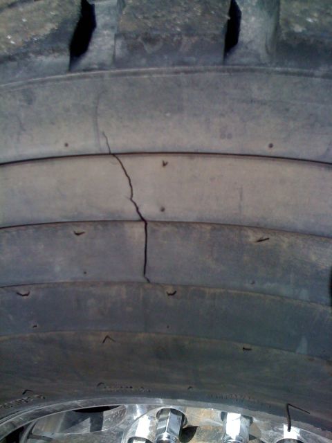 1996 Ford C-350 (Centurion Conversions) - Close-up of a light-truck tire's sidewall and shoulder showing a long vertical crack running across the smooth rib sections, with molded ru…