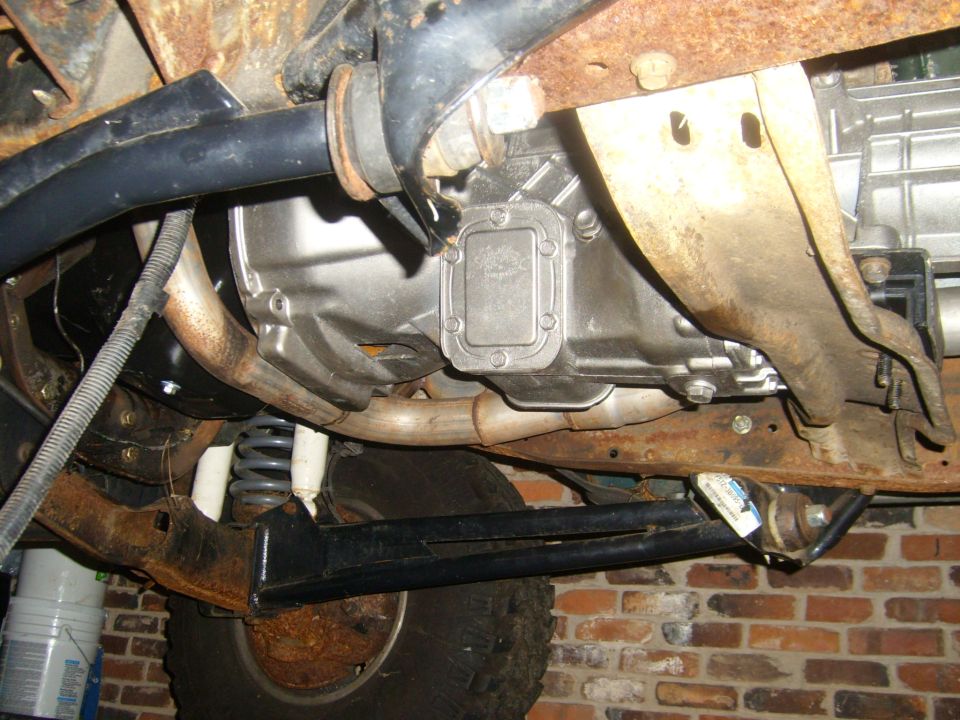 Undercarriage view showing transmission, exhaust pipes, and suspension parts. Brick wall in background