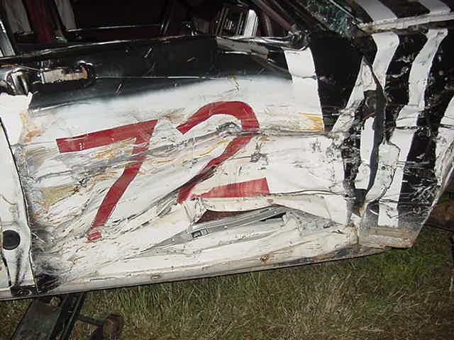 Crushed white race car door with red number 72, sitting on grass at night
