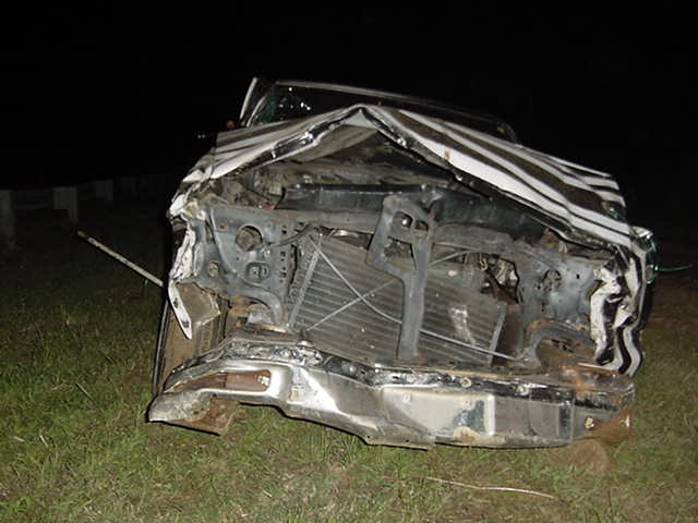 Wrecked silver car with crushed front end and exposed radiator at night in a field