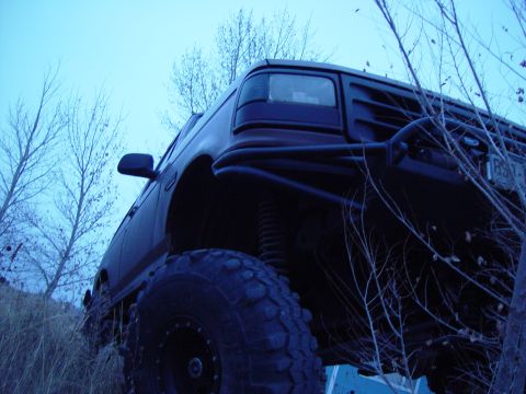 1993 Ford Bronco - This modified Ford truck features a custom front bumper and is parked on uneven terrain with bare trees in the background.