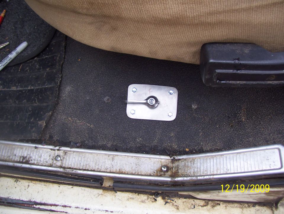 Interior floor mount with a silver lever and bolts, next to a tan seat and black carpet