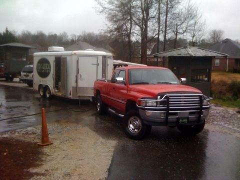 1989 Ford Bronco - A red Dodge Ram pickup truck is parked on a wet surface, towing a white trailer with a circular logo.