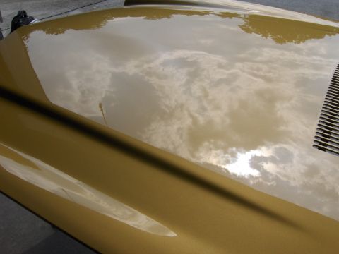 1971 Chevrolet Camaro - Sun-glossed gold metallic car hood and fender reflecting the cloudy sky, with a black cowl vent at the edge.