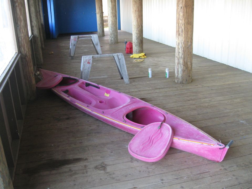 Pink kayak and paddle on wooden deck, with sawhorses and canisters in background