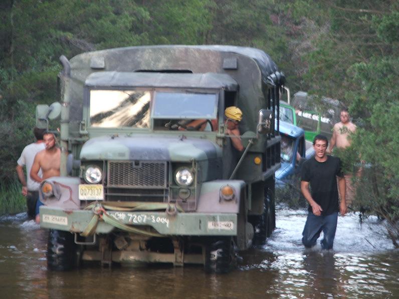 Camouflage military truck driving through water with people around