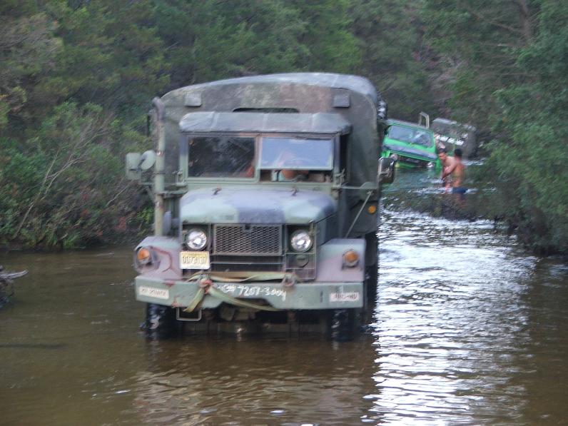 Front view of a camouflage military truck in water, with a green vehicle partially submerged behind it