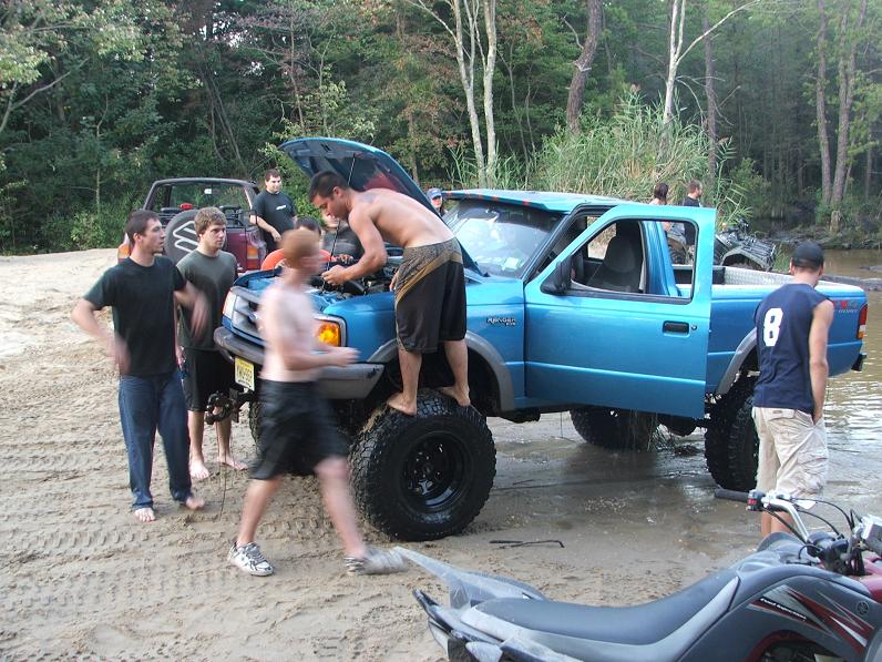 Blue pickup truck with hood open, people working on engine, lifted suspension, off-road tires, in shallow water