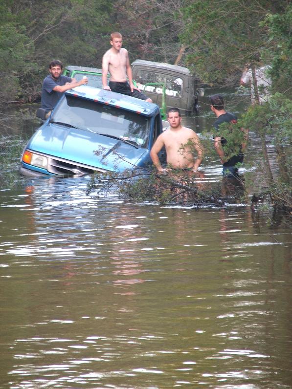 Light blue compact pickup swamped in deep water with people standing on it and wading nearby