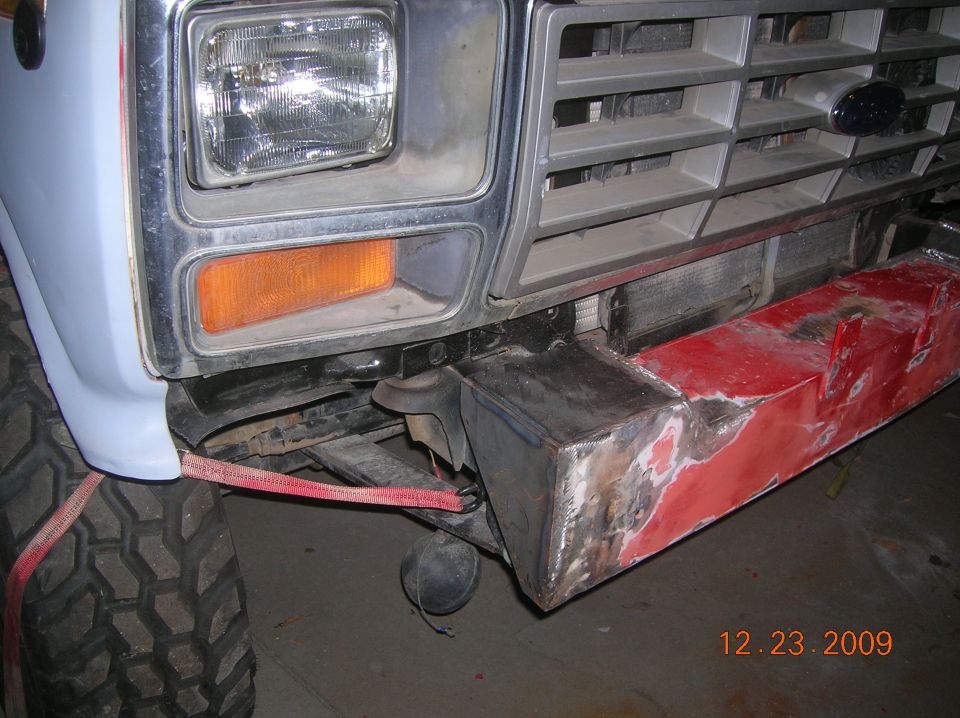 Front end of a Ford truck, likely 1980-1986, with a custom fabricated bumper. Red tow strap is attached to the tire