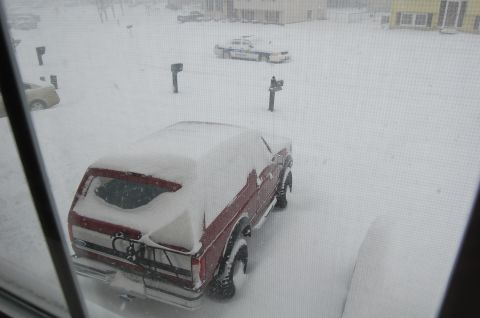 2002 BMW M3 - A red pickup truck is parked in a snow-covered driveway, with other vehicles and houses barely visible through the falling snow.