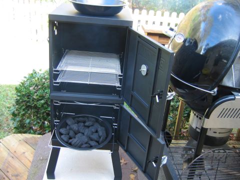 1992 Ford Bronco - A black vertical charcoal box smoker sits open on a backyard patio beside a black Weber kettle grill with a lit temperature gauge, a pan of…