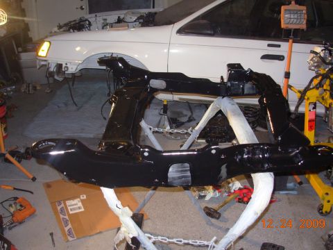 1991 Ford Taurus - A white car is partially disassembled in a garage, with a freshly painted black subframe in the foreground.