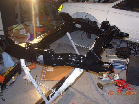 1991 Ford Taurus - A car subframe is painted black and displayed in a garage with a white vehicle behind it.