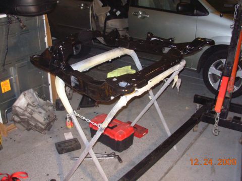 1991 Ford Taurus - Car subframe assembly on a stand, with a transmission nearby. A silver minivan is visible in the background.