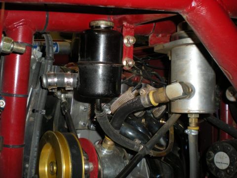 Suzuki Samurai - Detail of a hydraulic steering setup in a red tube-frame engine bay: a black remote reservoir with a knurled cap is bracketed to the frame…