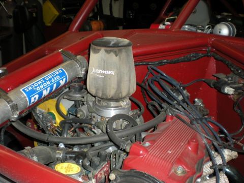 Suzuki Samurai - Engine bay of a red off-road build showing a conical air filter wrapped in an Outerwears pre-filter mounted on the throttle body, next to a…