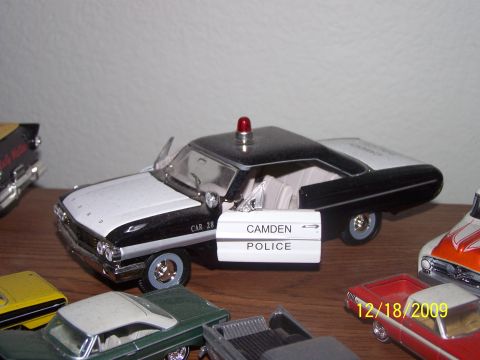 1972 Ford Ranchero - A collection of model cars is displayed on a wooden surface, including a black and white police car with an open door.