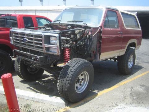 1986 Ford Bronco - A two-tone red and tan Ford Bronco SUV mid-build, viewed from the front three-quarter angle, with front fenders, hood, and grille component…