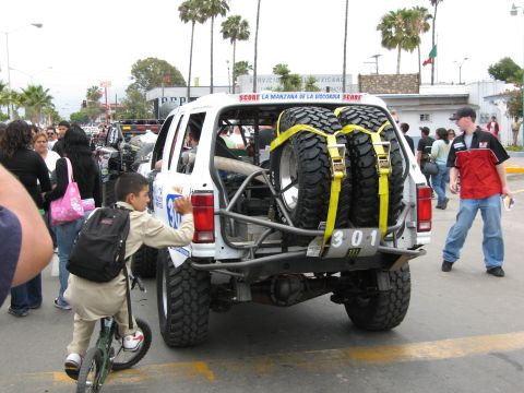 1986 Ford Bronco - A white off-road truck with spare tires is parked on a street, with people and palm trees in the background.