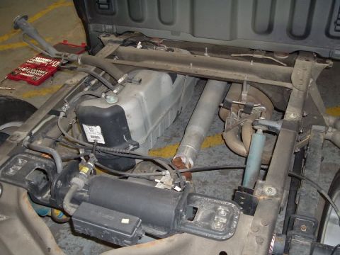 2007 Ford Mustang - Close-up of a vehicle's front-end components with the hood raised, showing a light-colored plastic reservoir/canister, black cylindrical co…