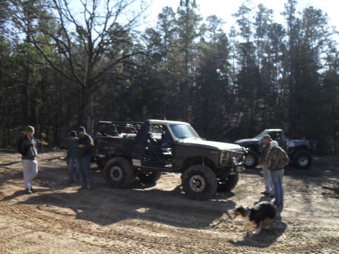 1986 Ford Bronco - A heavily modified SUV with a lifted suspension and large off-road tires is parked in a wooded area with other vehicles present.