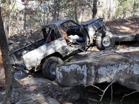 1986 Ford Bronco - An off-road truck navigates a rocky terrain with its driver's side door open and a roll cage installed.