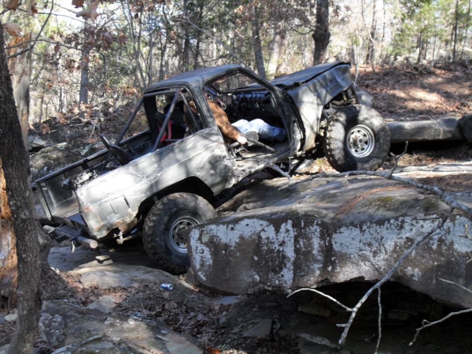 Damaged off-road truck on rocks, driver's side door open, roll cage visible