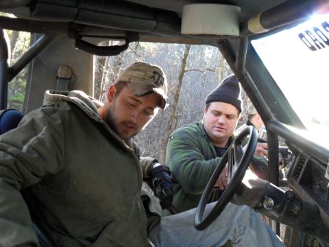 1986 Ford Bronco - Two men are inside a vehicle with a roll cage, possibly preparing for an off-road adventure.
