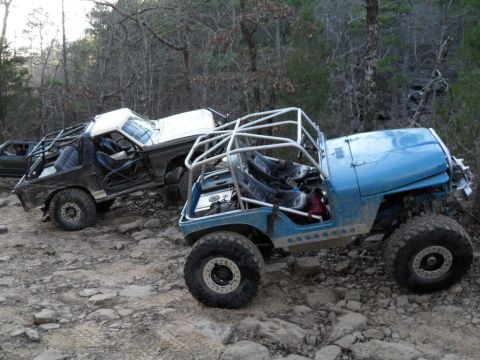 1986 Ford Bronco - Two modified off-road vehicles, a 1986 Ford Bronco and a blue Jeep, navigate a rocky trail in a wooded area.