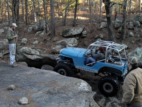 1986 Ford Bronco - Off-road Jeep Wrangler with custom roll cage tackles a rocky obstacle, observed by two people.