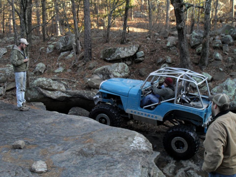 Blue Jeep Wrangler with roll cage navigating rocky trail, two men watching